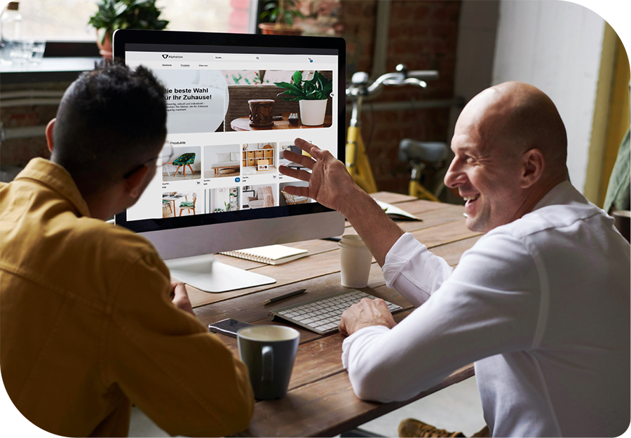 Zwei Männer sitzen an einem Tisch vor einem Monitor. Auf dem Monitor ist ein Webshop zu sehen. Der rechte Mann lacht und gestikuliert freundlich.