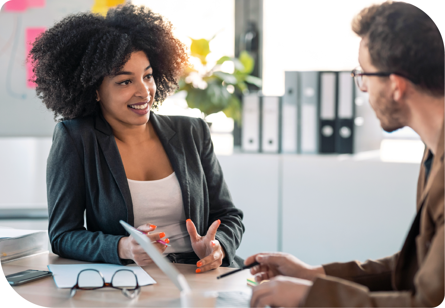 Eine junge Frau mit lockigem Haar in einem professionellen Gespräch mit einem Mann. Beide sitzen an einem Schreibtisch in einem modernen Büro, umgeben von Aktenordnern und Dokumenten. Die Frau lächelt und gestikuliert freundlich.